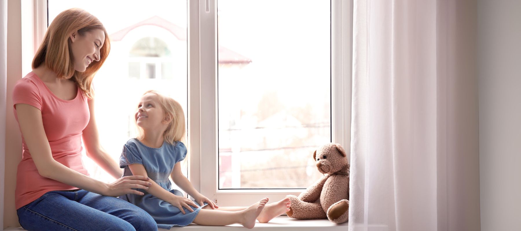 Mutter und Tochter sitzen fröhlich auf der Fensterbank neben hellen Vorhängen im Kinderzimmer.